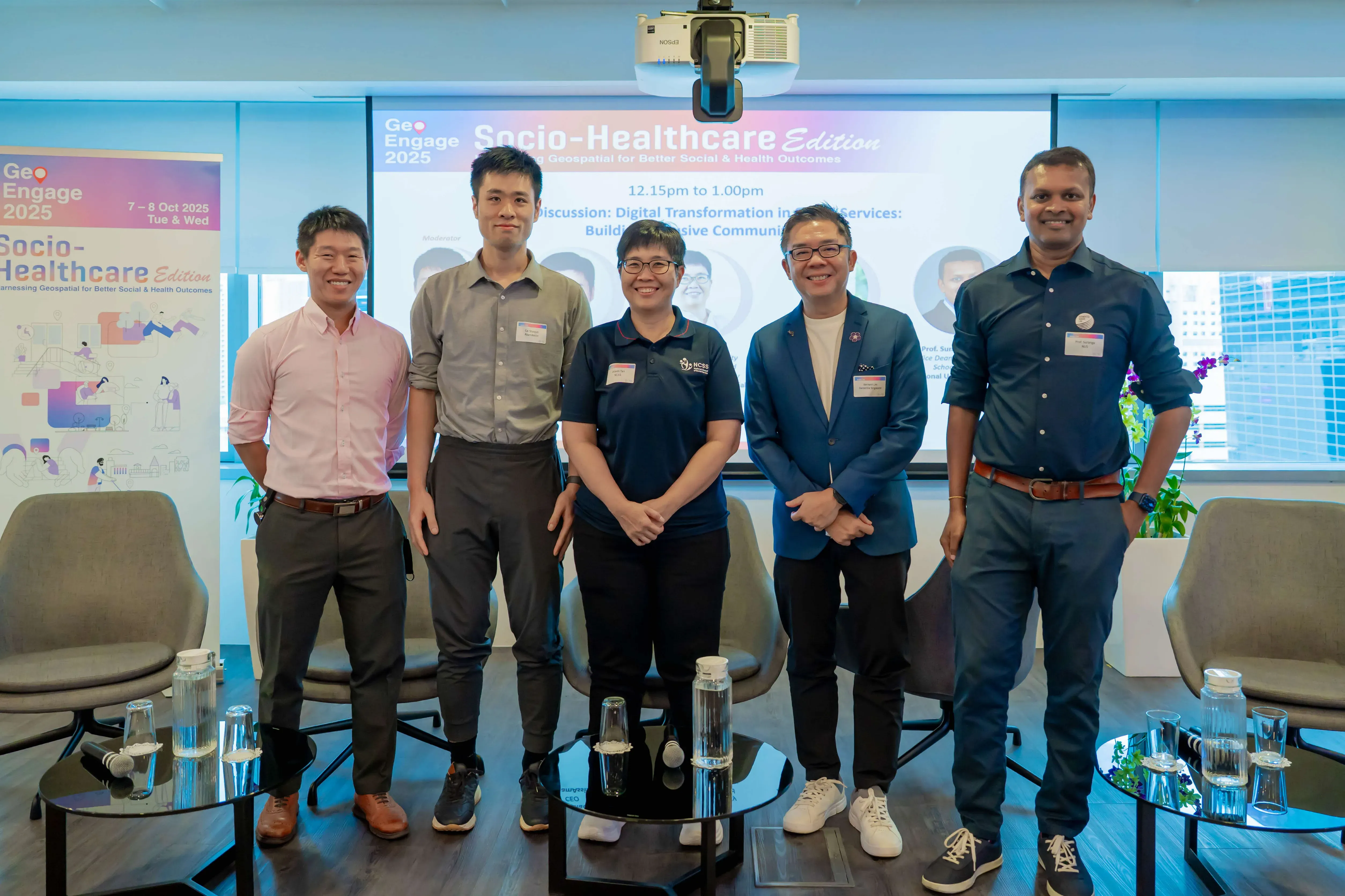 Five people stand smiling at a GeoEngage 2025 Socio-Healthcare Edition panel event with a projected screen behind them.